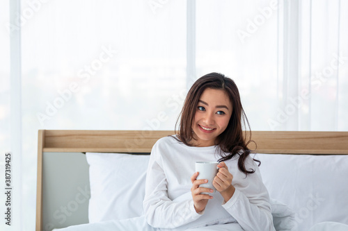 Young woman relaxing and drinking cup of coffee or tea on a cold winter day in the bedroom. Casual beautiful Asian woman sitting on the bed with cup of coffee in the house.