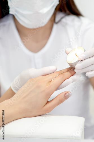 Manicure master applying warm wax from candle on fingernails of young woman in nail salon