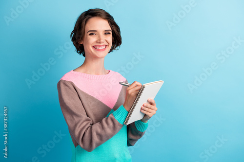 Photo portrait of smiling cheerful female student taking writing essay in diary with pen isolated on vivid blue color background
