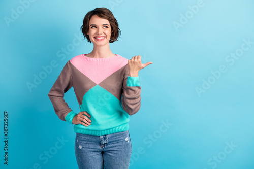 Photo portrait of woman pointing thumb looking at blank space with one hand on waist isolated on pastel light blue colored background