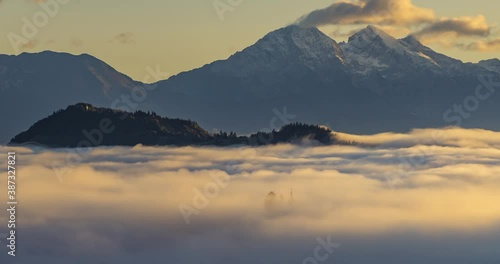Time lapse low altitude inversion clouds rolling in Alpine valley, Slovenia. Saint Thomas church tower visible above the fog. Alpine mountains in background. Right truck parallel moving, wide angle