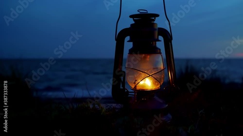 Old classic oil lantern burning with an orange flame by the ocean at dusk. Blurred background.