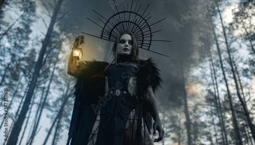 Fotografija Woman in image of witch stands with glowing lamp against black smoke in forest