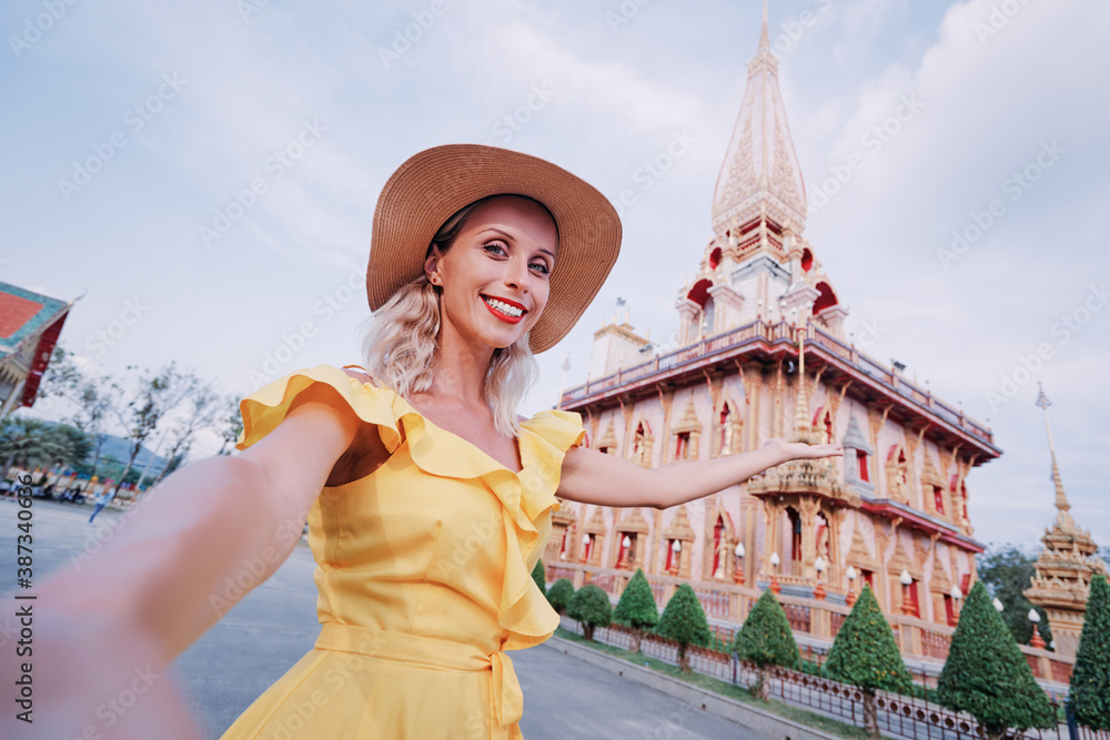 Travel by Asia. Young woman in hat and yellow dress taking selfie on smartphone near Chalong buddhist temple on Phuket Island in Thailand.
