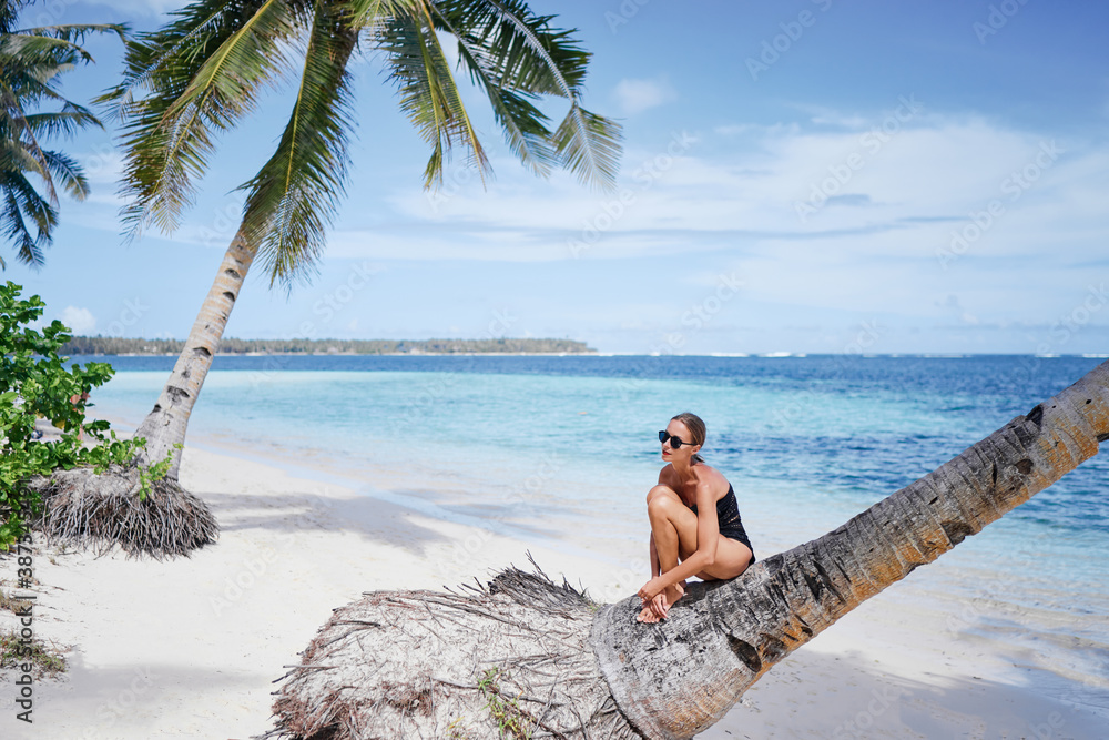 Vacation on the seashore.Young woman in black swimsuit on the beautiful tropical beach sitting on the palm tree.