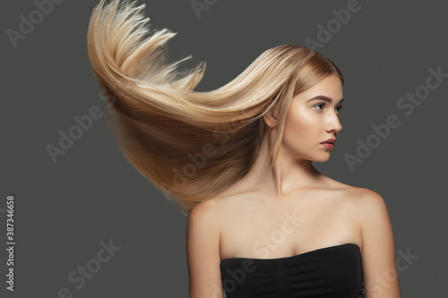 Light. Beautiful model with long smooth, flying blonde hair on dark grey studio background. Young caucasian model with well-kept skin and hair blowing on air. Concept of salon care, beauty, fashion.