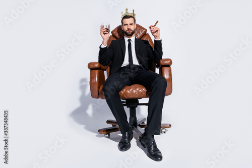 Full body low below angle view photo of handsome business guy sit chair hold rum glass cuban cigar chief wear crown black blazer pants tie shirt shoes suit isolated grey background photo