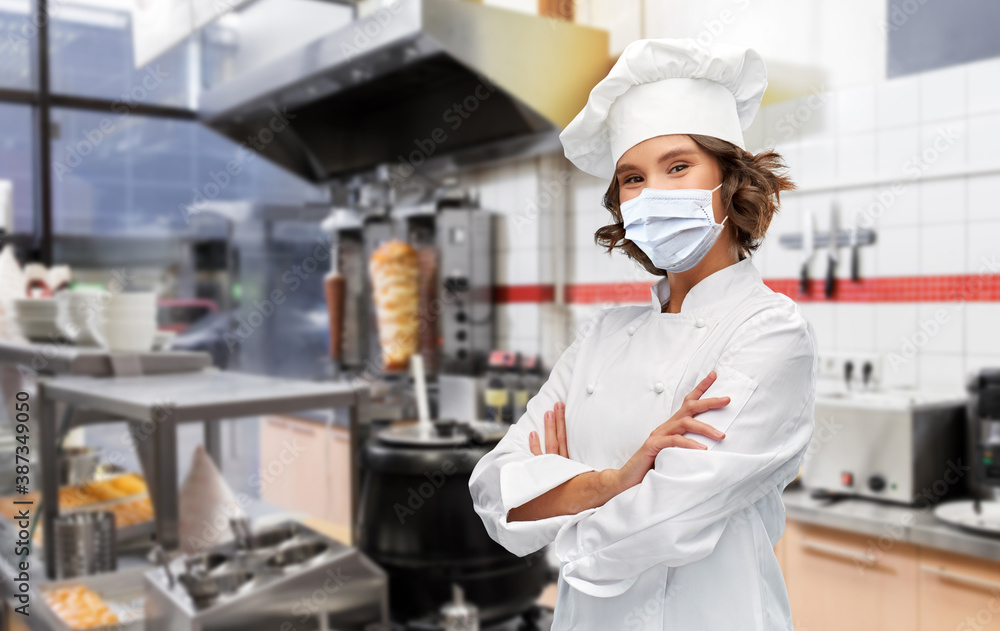 cooking, culinary and health concept - female chef in toque wearing face protective medical mask for protection from virus disease with crossed arms over kebab shop kitchen background