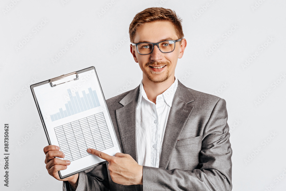 Young handsome brunet man in glasses in suit smiles and points at business with documents and graphs on white isolated background Young handsome brunet man in glasses in suit smiles and points at business with documents and graphs on white isolated background