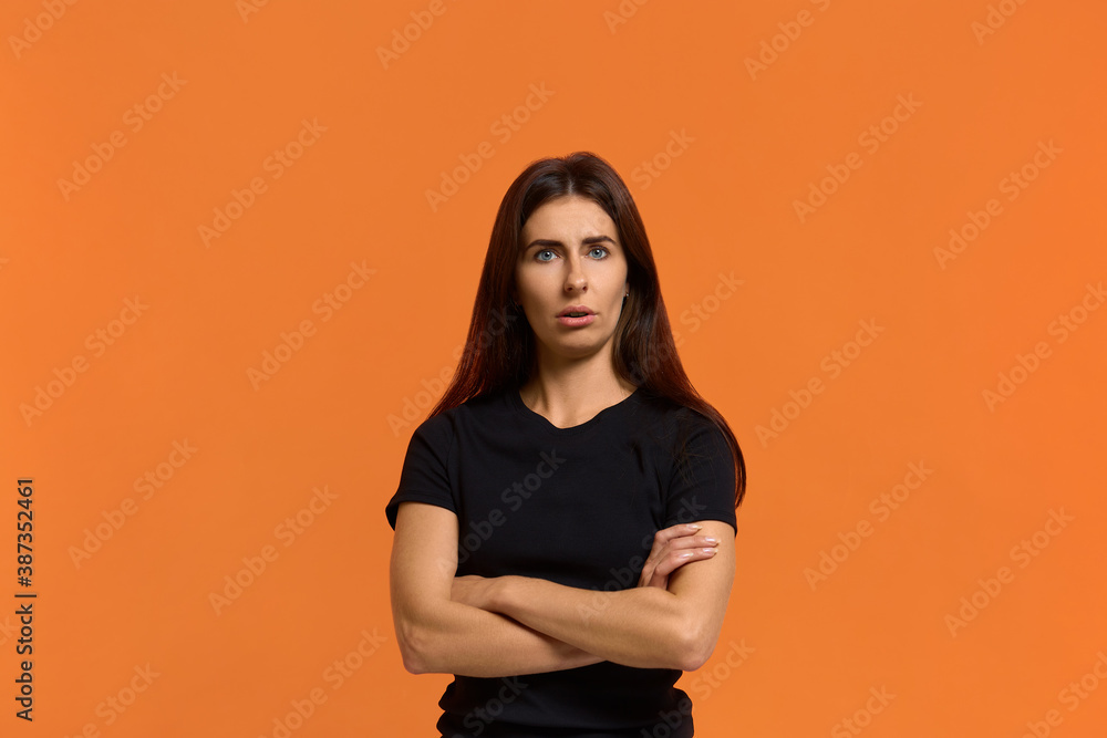 Obraz premium Are you serious. Portrait of attractive funny emotional caucasian woman in black t-shirt looking at camera, keeping arms crossed and open mouth, feeling shocked. Isolated over an orange wall