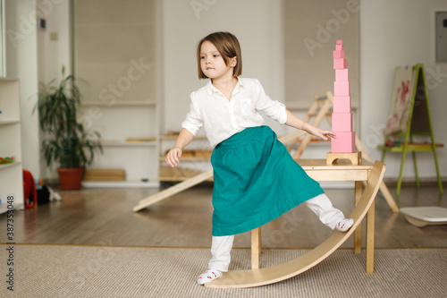 child girl in a linen apron sways on balancing board, developing sensory and sports activities in montessori and earlier child development, children's independence