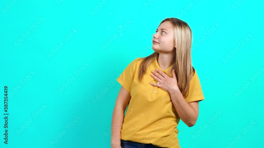 The modest young woman looks around and finally raises her hands disapprovingly. Beautiful Caucasian woman in yellow T-shirt and long blond hair standing on blue background. A little fat teenager The modest young woman looks around and finally raises her hands disapprovingly. Beautiful Caucasian woman in yellow T-shirt and long blond hair standing on blue background. A little fat teenager