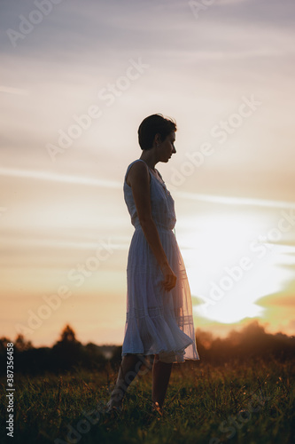 Girl in a white dress walks in the field during sunset. © Evgenii Starkov