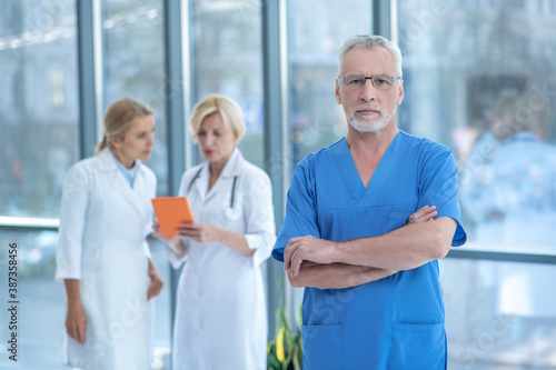 Male doctor looking with folded arms into the camera, female colleagues studying something on tablet behind