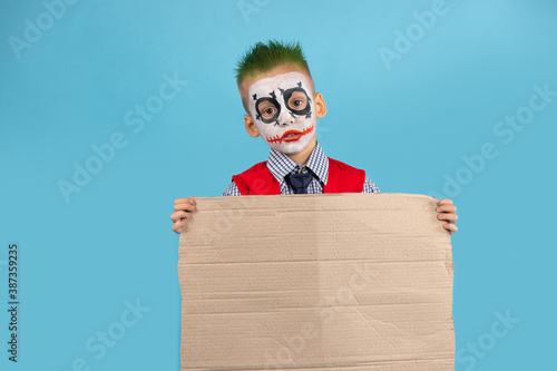 Sad Halloween preschooler with face art. Child holding blank protest cardboard isolated on blue background. Social problems and people concept. Preparing for Halloween. photo