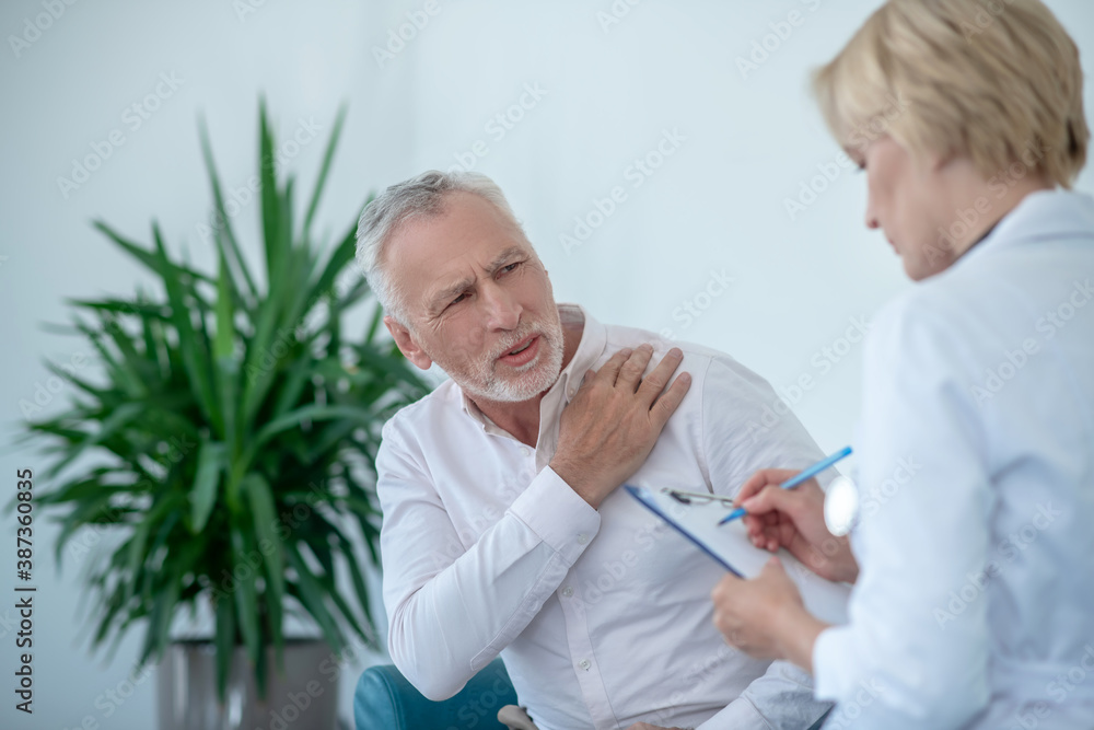 Fototapeta premium Gray-haired male patient suffering from shoulder pain, female doctor taking notes