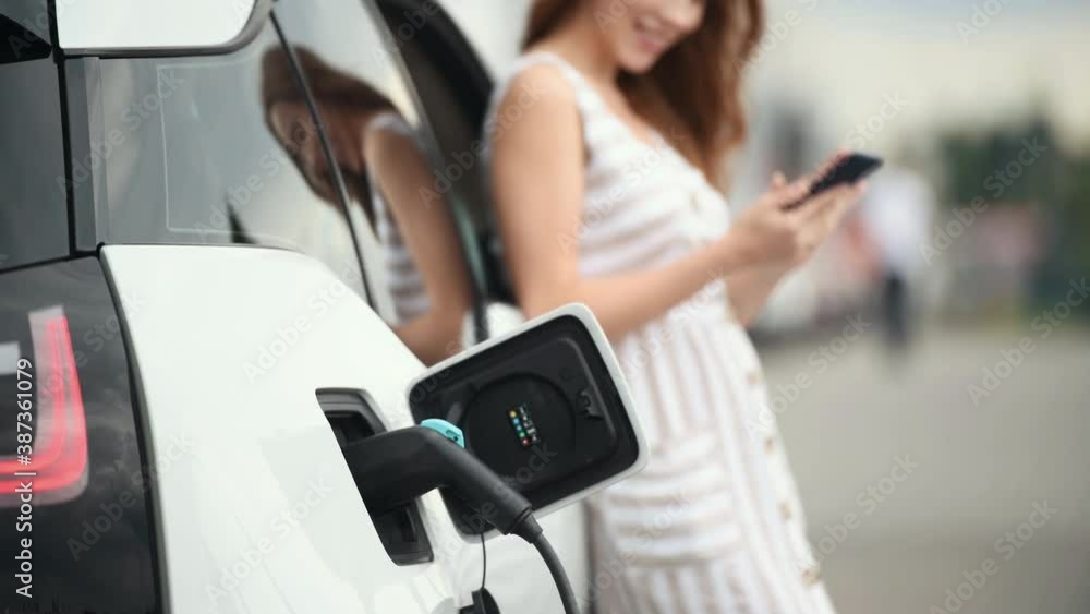 Woman stands with phone near her electric car and waits when vehicle will charged. Woman stands with phone near her electric car and waits when vehicle will charged.