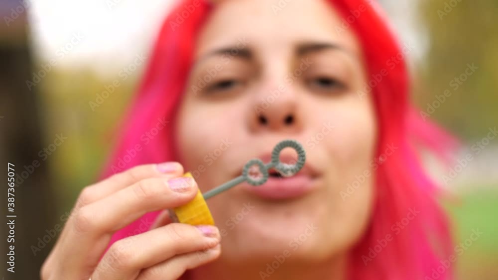 Happy adult girl blowing soap bubbles front camera in nature. Close-up. Good-looking girl blows bubbles at camera and has fun. Portrait young cheerful caucasian woman with bright pink hair. Happy adult girl blowing soap bubbles front camera in nature. Close-up. Good-looking girl blows bubbles at camera and has fun. Portrait young cheerful caucasian woman with bright pink hair.