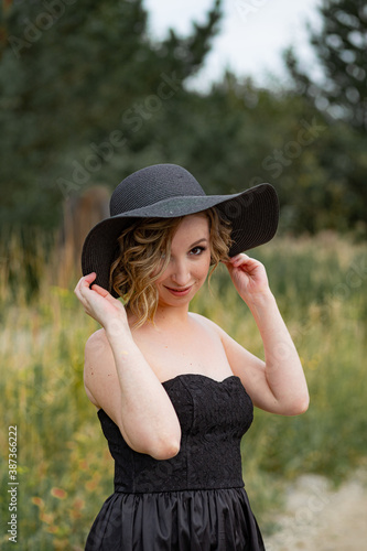 An attractive girl with a short haircut in a black dress and a black hat stands in nature. Hats fashion. A close up portrait of a charming young woman in a fashionable black hat 