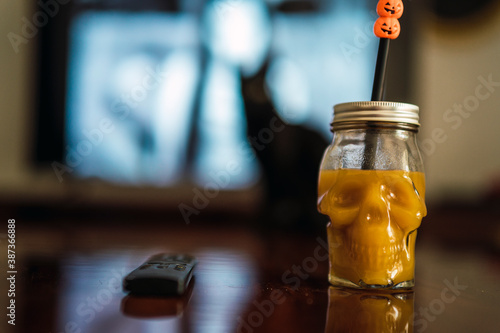 Vaso con forma de calavera rellena de liquido naranja sobre la mesa con gato negro de fondo