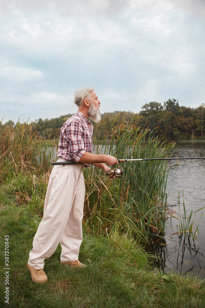 Vertical full length shot of a senior man fishing with a rod on countryside lake Vertical full length shot of a senior man fishing with a rod on countryside lake