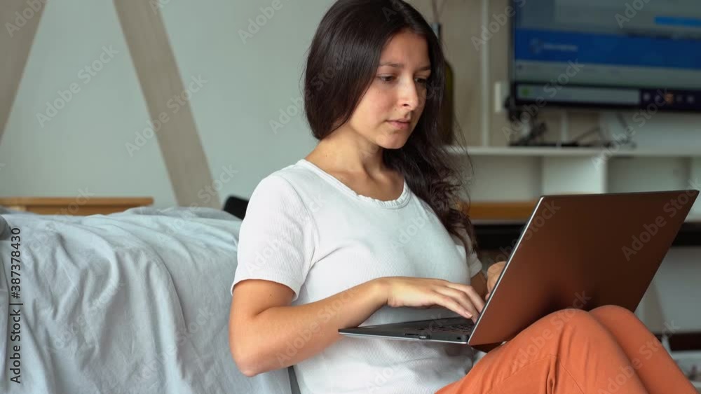 Beautiful young adult girl sitting on the floor by the bed early in the morning and using her laptop, freelance and remote working concept Beautiful young adult girl sitting on the floor by the bed early in the morning and using her laptop, freelance and remote working concept