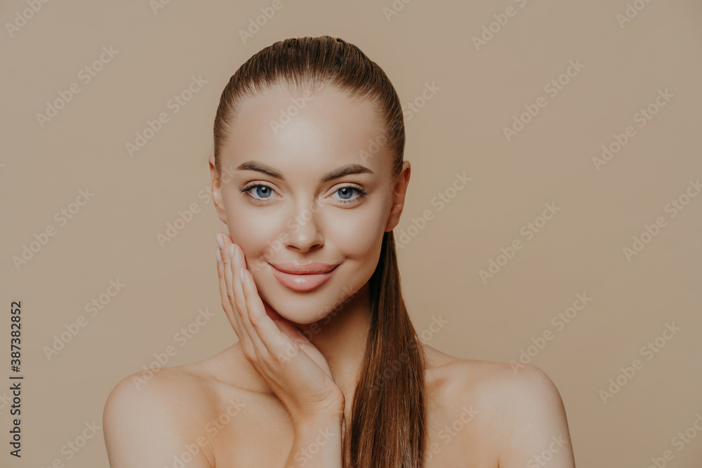 Photo of lovely young woman has smooth healthy skin after beauty procedures and receiving facial massage, stands bare shoulders, has long dark straight hair, isolated over beige wall cares of her body Photo of lovely young woman has smooth healthy skin after beauty procedures and receiving facial massage, stands bare shoulders, has long dark straight hair, isolated over beige wall cares of her body