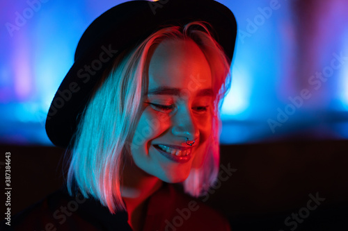 Hipster woman with blond hairstyle standing on neon glowing background. City at night. Hat, nose piercing. Beautiful attractive girl.