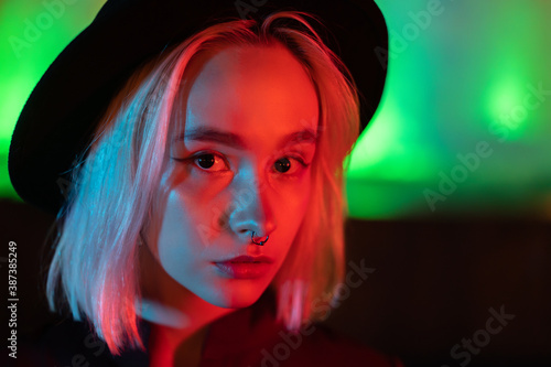 Hipster woman with blond hairstyle standing on neon glowing background. City at night. Hat  nose piercing. Beautiful attractive girl.