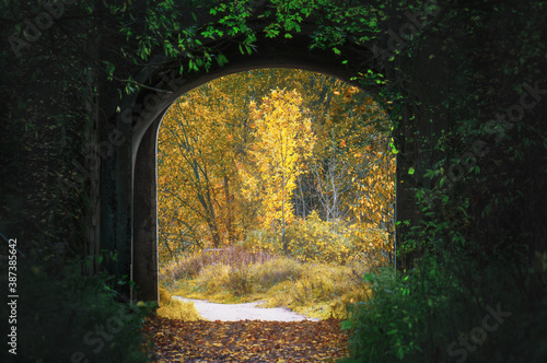 Gate to autumn: an ancient abandoned arch, green foliage in front of it, and an autumn yellow landscape behind.