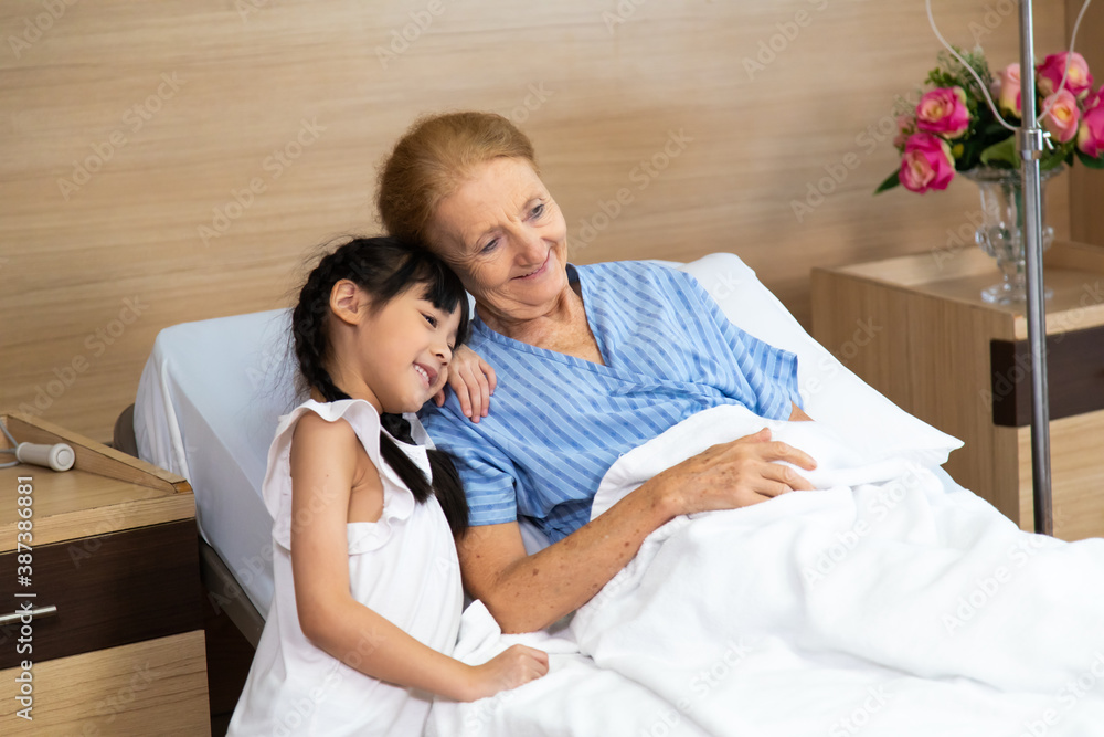 Fototapeta premium Happy elderly patient lying on hospital bed with grandchild visitor. grandmother and granddaughter enjoying talking together.