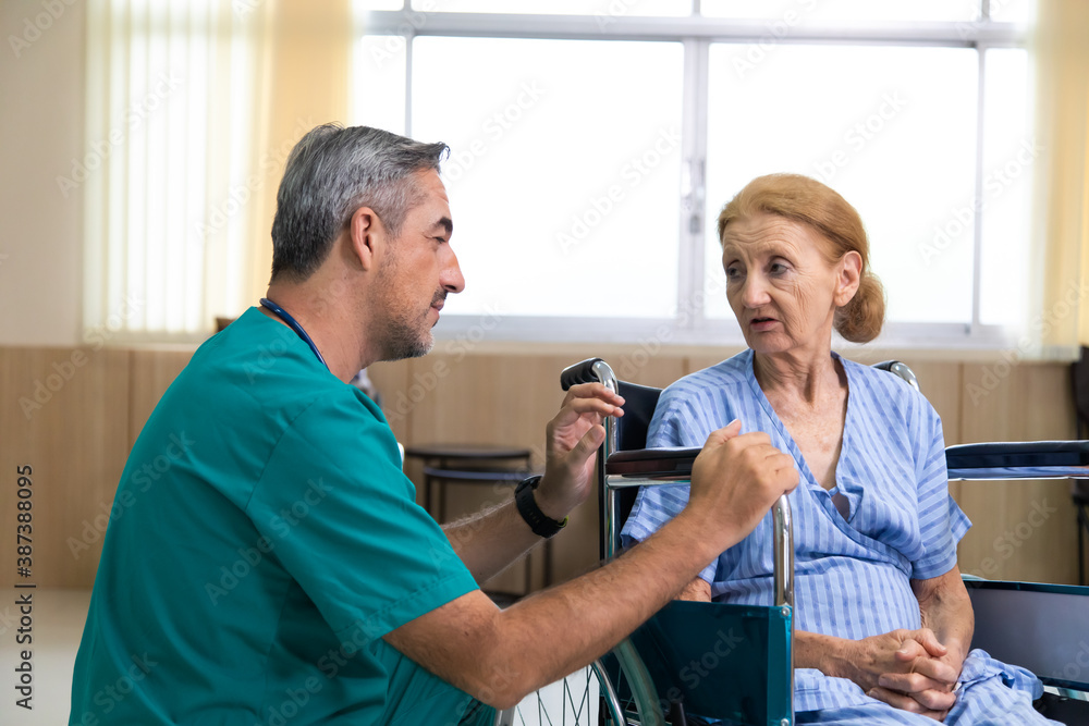 Middle age surgeon talking and consultant to senior woman patient in wheelchair. Senior woman on wheelchair and her young caregiver talk care at each other. Middle age surgeon talking and consultant to senior woman patient in wheelchair. Senior woman on wheelchair and her young caregiver talk care at each other.