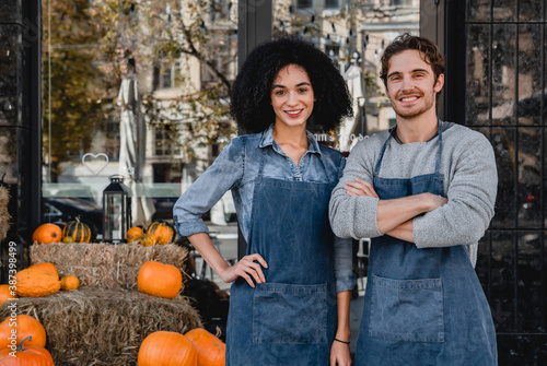 Portrait of young man and woman cafe owners standing outdoor front of their cafe with decorating pumpkins preparing for halloween.