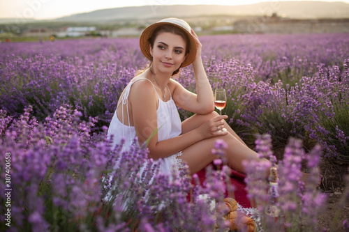 Portrait of pretty girl is wearing big hat having picnic and drinking wine in lavender field, France.