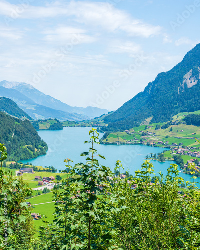 Panorama d'un lac bleu turquoise avec un petit village encastré entre des montagnes, avec de la végétation au premier plan, un jour ensoleillé avec des nuages