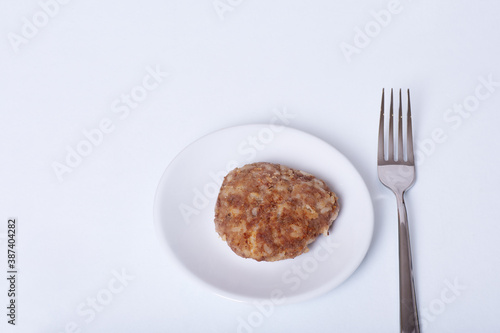 Cooked meat patty on a white plate next to a fork isolated on white background  close-up  copy space.