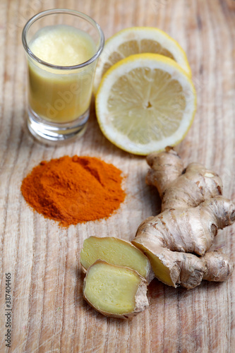 Ginger drink or ginger shot and healthy ingredients like ginger root, lemon, turmeric on wooden background.