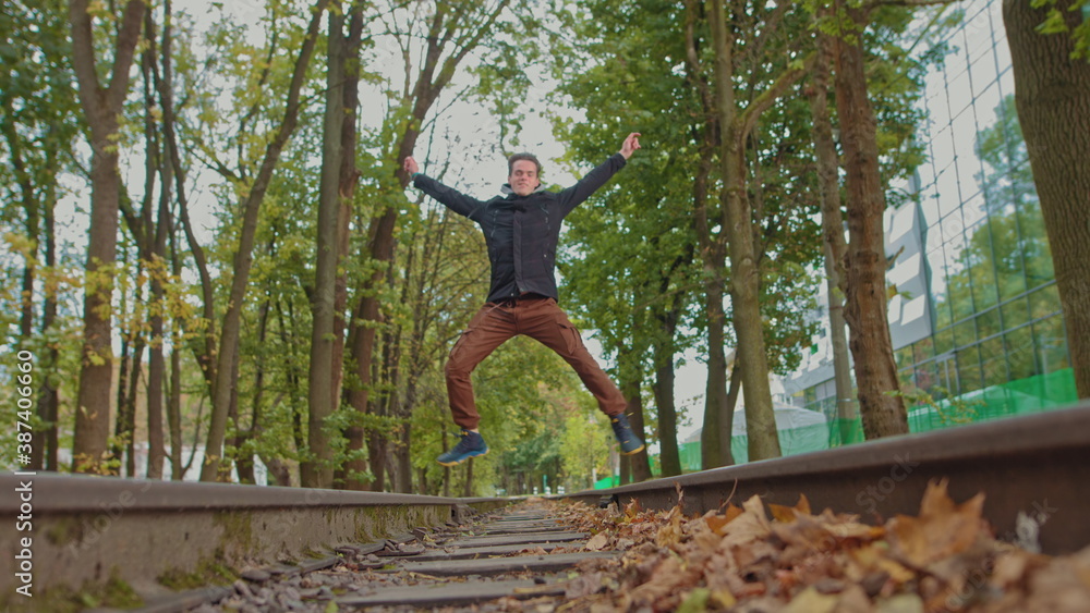 Naklejka premium Playful happy man teenager, student, rejoices at success. A young guy funny bouncing in the park and jumps with happiness against the backdrop of a beautiful sunset. Slow motion. on the front