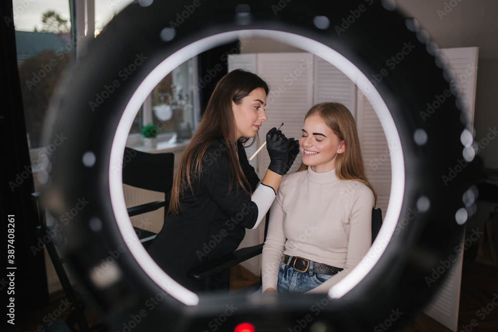 Fototapeta premium Brow master applies henna on eyebrows for beautiful blond hair woman. Photo with ring light