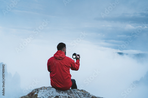 Young bearded hiker wearing red jacket taking photos at mountain with fog and mist surrounding peaks and forest