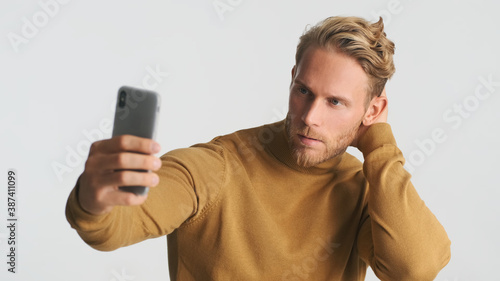 Handsome stylish bearded man wear turtleneck looking confident taking selfie on smartphone over white background. Male model posing in studio
