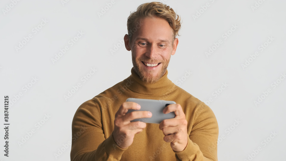 Attractive excited bearded man look happy playing in game on smartphone and smiling over white background
