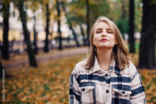 A white girl in a plaid shirt stands in an autumn Park, her head up and her eyes closed. Listens and enjoys nature. Human emotions, pleasure. Space for text.
