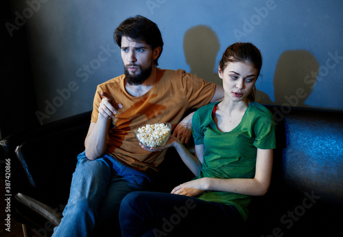 A woman and a man with popcorn on a leather sofa indoors watching TV in the evening