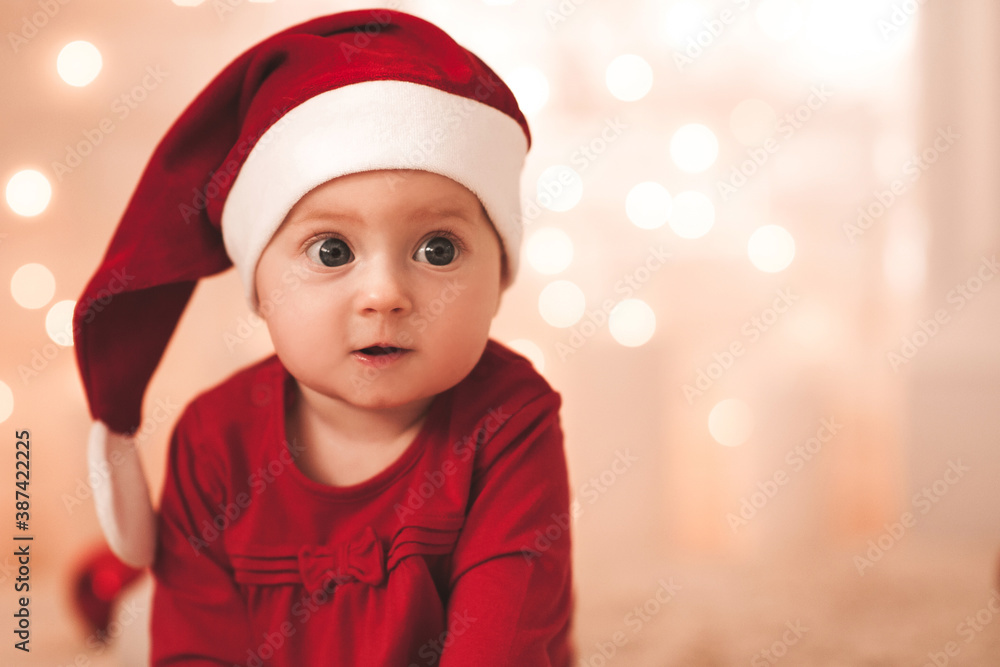 Cute baby girl 1 year old wearing red santa hat and dress over glowing Christmas lights in room closeup. Winter holiday season. Childhood. Cute baby girl 1 year old wearing red santa hat and dress over glowing Christmas lights in room closeup. Winter holiday season. Childhood.