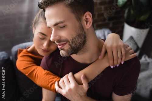 young man with closed eyes embraced by tender girlfriend on blurred background