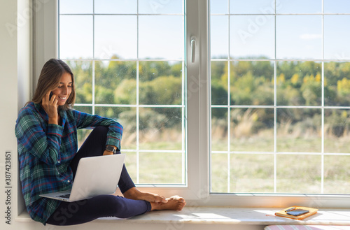 Portrait of young smiling businesswoman wearing pajama and calling her best friend, having break, telling something funny, sitting n the windowsill
