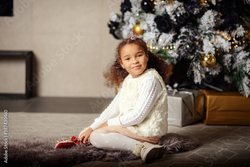 Happy smiling mixed girl with dark hair sitting on the carpet. Christmas tree in the background. © lialia699