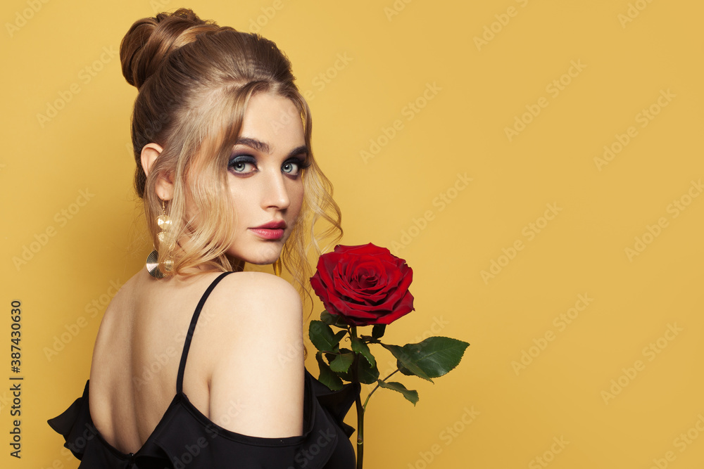 Fashion beauty portrait of beautiful woman with blonde hairdo and makeup holds red rose flower on black background