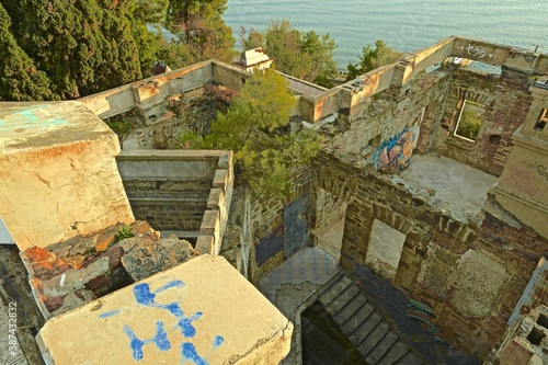 Top view of the ruins of stone walls and the broken roof of an old abandoned building in the classical style, surrounded by southern vegetation, against the background of the sea